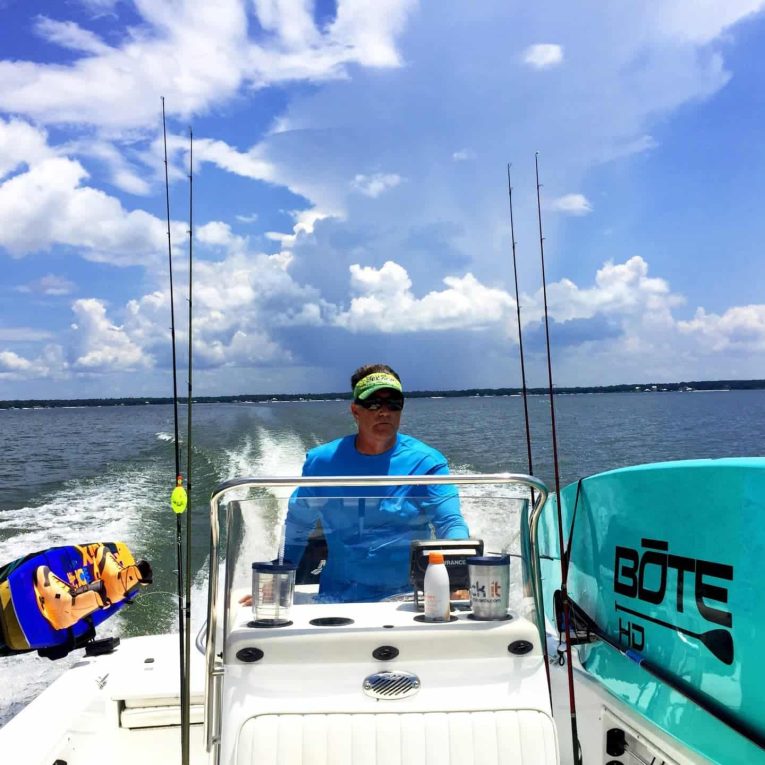 A man controls a boat while transporting a blue wakeboard and a light blue paddleboard on its side.