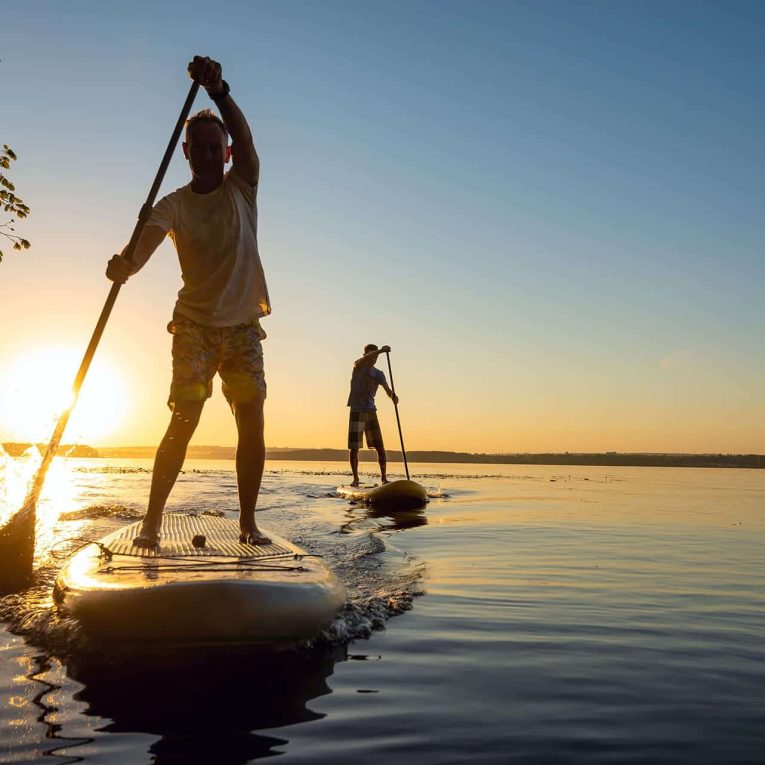 Two men paddleboarding on calm waters with sunset view in the background.