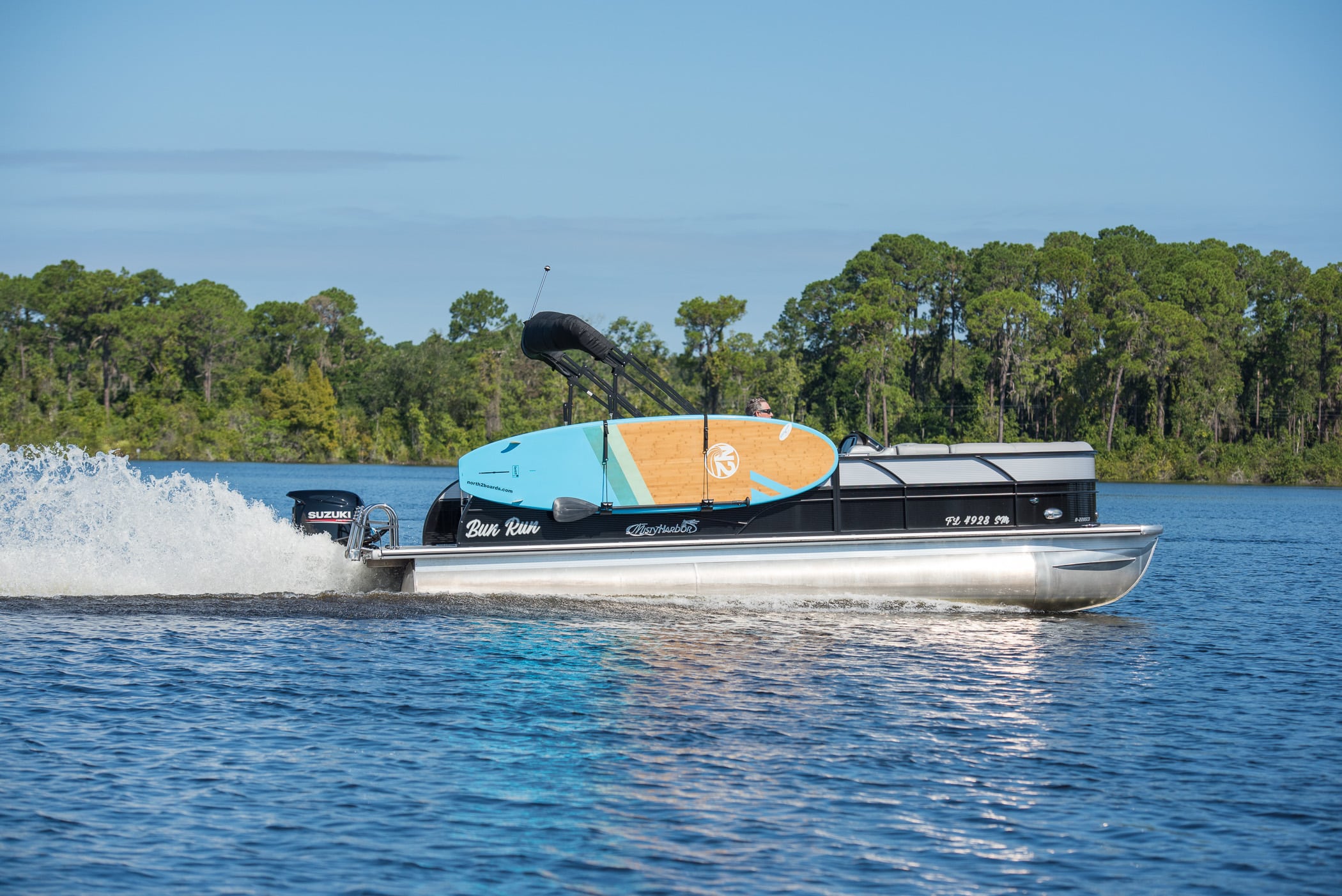 Pontoon boat on the water with a Paddleboard stored in the PS1 Paddleboard Storage Rack for Pontoon Boats, on the side.